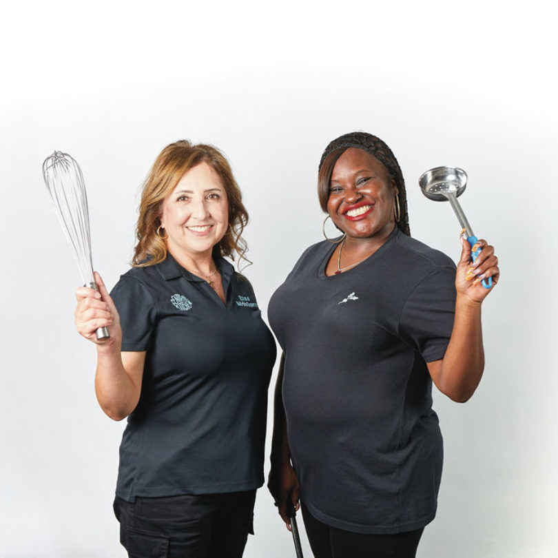 Two Central Kitchen team members smiling and standing side by side against a white background, holding a whisk and a ladle as they pose proudly in their black work shirts.