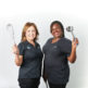 Two Central Kitchen team members smiling and standing side by side against a white background, holding a whisk and a ladle as they pose proudly in their black work shirts.