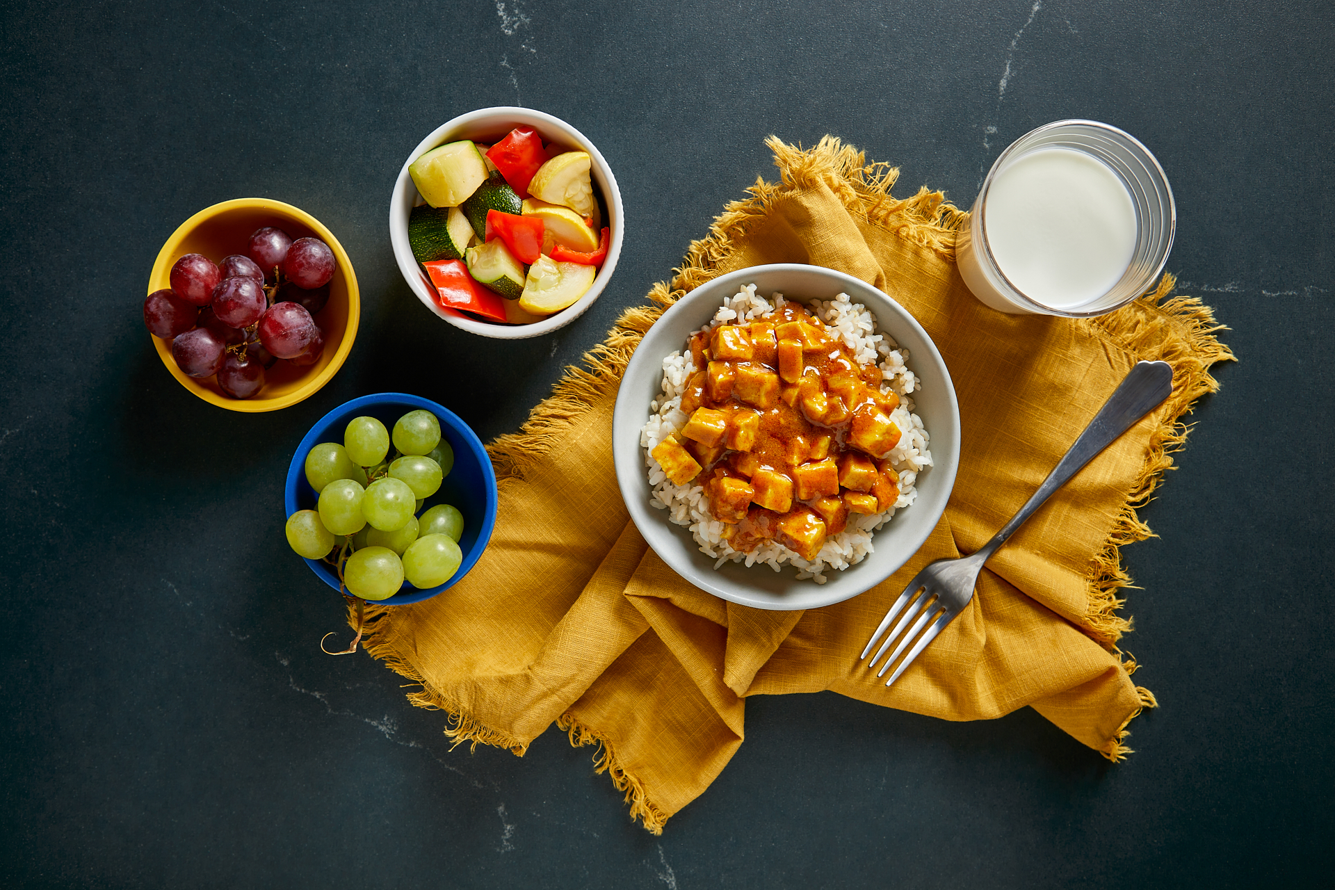 Overhead photo of yellow napkin with bowl of rice and curry tofu, side ides of grapes and vegetables and a glass of milk.