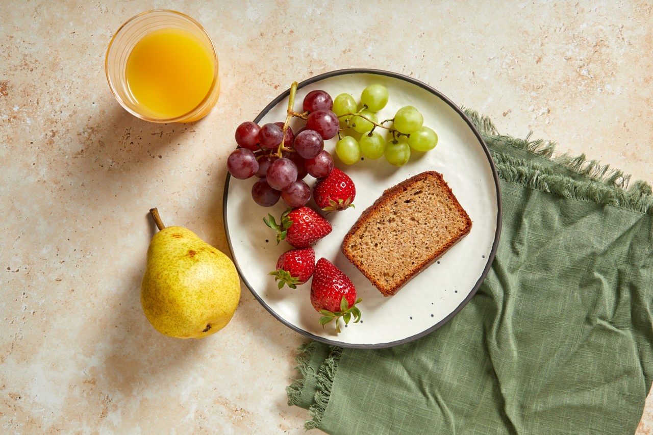 Header image of a plated breakfast including a slice of spice bread, grapes, strawberries, a whole pear and a glass of orange juice set on top of a green table linen.