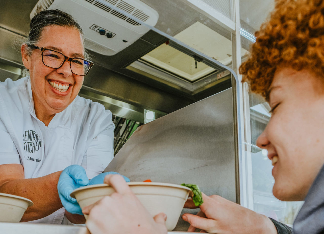 Smiling woman in a white chefs coat with the Central Kitchen logo on the pocket is handing a bowl of food through a food truck window to a smiling teenage boy with red curly hair